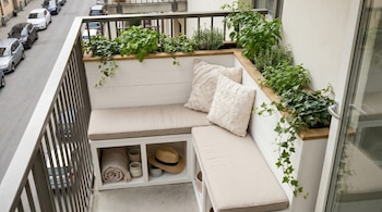 Balcón con banco blanco y cojines beige, jardineras con plantas, almohadas. En el almacenamiento inferior hay una manta, tazas y un sombrero. Calle con coches.