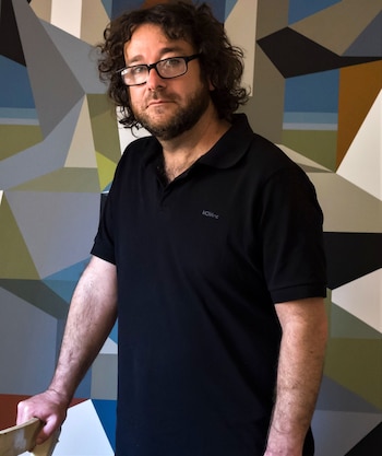 Hombre con cabello rizado y gafas, barba, camiseta polo negra, parado frente a una pared con un patrón geométrico abstracto multicolor
