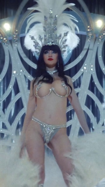 Una mujer lleva un traje plateado de vedette con plumas en la cabeza y pedrería delante de una estructura decorativa iluminada