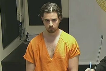 Hombre de cabello oscuro y barba con un uniforme naranja en una sala con un micrófono y fondo blanco