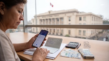 Mujer sosteniendo un teléfono inteligente junto a una libreta de ahorros, una calculadora y billetes de cien soles sobre un escritorio.