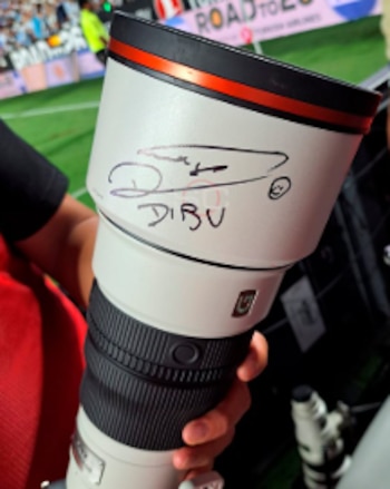 Primer plano de un teleobjetivo blanco con una firma en marcador negro y el nombre 'DIBU' escrito. Se ve una mano sosteniendo el lente en un estadio