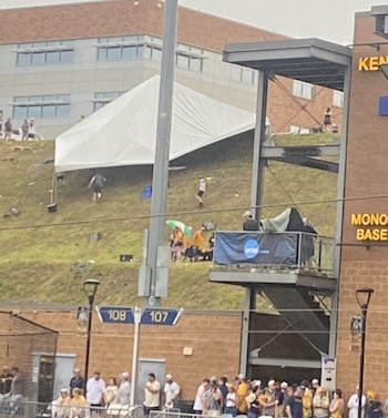 Tienda de campaña blanca parcialmente colapsada en una ladera cubierta de hierba, con personas dispersas y parte de un estadio de béisbol con público