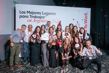 Grupo de diecisiete personas sonrientes, hombres y mujeres, posando frente a un cartel "Los Mejores Lugares para Trabajar™". Cae confeti rojo sobre ellos