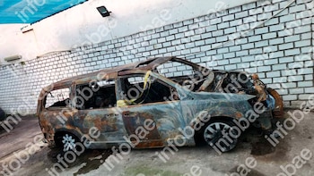 Automóvil familiar gris oscuro quemado, con óxido y hollín, estacionado junto a una pared de ladrillos blancos y un suelo de cemento