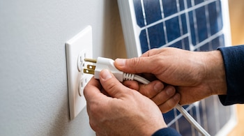 Manos sostienen un enchufe blanco de tres patas frente a un tomacorriente de pared, con un panel solar azul en el fondo.