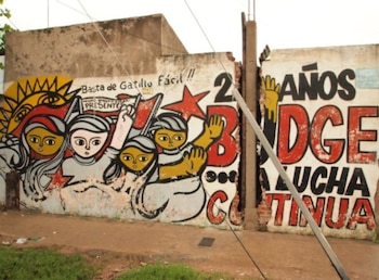Mural con figuras estilizadas, sol, y texto "Basta de Gatillo Fácil" y "25 AÑOS LUCHA CONTINUA" en una pared de hormigón con cables y poste