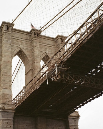 Vista de cerca de la torre de piedra del Puente de Brooklyn, mostrando sus arcos, los cables de suspensión, la estructura de celosía de la calzada y la bandera de EE.UU. en la cima