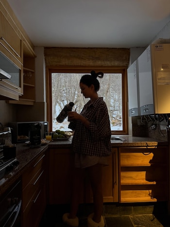 Una mujer de perfil prepara mate en una cocina con ventanas. Se ven electrodomésticos, frutas, una bebida y un paisaje invernal nevado