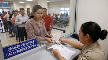 Una mujer entrega billetes de dólar a una empleada en una ventanilla con el letrero "Caja - Tarifas de Visa". Una fila de personas espera detrás.