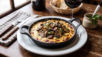 Una sartén pequeña sobre un plato blanco contiene queso provoleta y hongos, junto a cubiertos, pan y una copa de vino en una mesa.