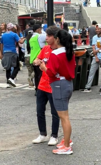 Un hombre y una mujer con prendas rojas conversan de pie en la explanada de un estadio rodeados de aficionados
