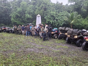 Hombres y cuatrimotos con equipaje agrupados en un terreno con barro frente a un obelisco blanco rodeado de vegetación