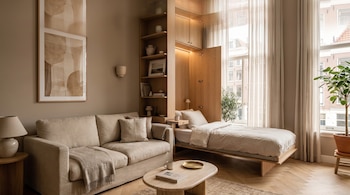 Un sofá beige junto a un mueble de madera que integra una cama desplegada y una mesa ovalada en una habitación iluminada.