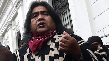 Un hombre indígena, Arturo Huenchullán, con cabello oscuro largo, poncho mapuche y bandana roja, habla frente a un edificio clásico blanco