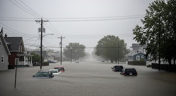 Calle residencial en una ciudad del norte de Estados Unidos totalmente inundada. Autos y casas parcialmente sumergidos bajo un cielo gris y lluvia intensa.