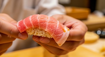 Primer plano de dos manos sosteniendo un trozo de chutoro nigiri, mostrando el arroz blanco debajo del pescado de color rosa marmoleado y cortado.