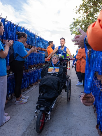 Un hombre empuja una silla de ruedas de tres ruedas ocupada por una mujer, entre voluntarios que aplauden y paneles llenos de medallas