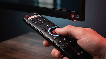 Una mano sostiene un control remoto negro frente a un televisor LG con un indicador luminoso rojo.