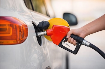 Primer plano de una mano sosteniendo una boquilla de gasolina roja y negra insertada en la boca del tanque de combustible de un automóvil blanco.