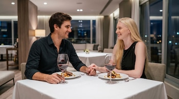 Un hombre y una mujer sentados en una mesa con mantel blanco. Cada uno tiene un plato con comida, un vaso de vino tinto y se toman de las manos.