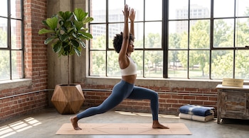 Una mujer con ropa deportiva realiza una postura de yoga sobre un tapete junto a grandes ventanas y una planta.