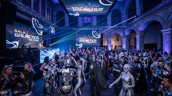 Personas disfrazadas de robots y alienígenas bailan bajo luces láser frente a un escenario con pantallas y un DJ.