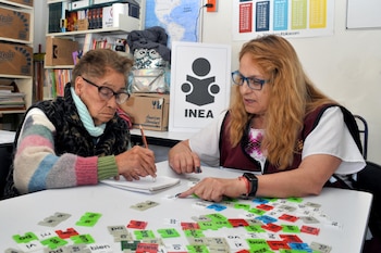 Dos mujeres en una mesa blanca con fichas de sílabas. La mujer mayor escribe en un cuaderno, la instructora señala las fichas. Lejano: un cartel de INEA y estantes