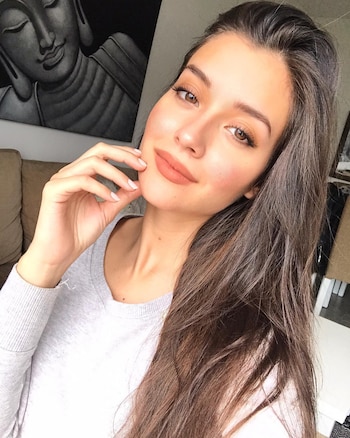 Manuela Lori, mujer joven con cabello castaño oscuro, suéter gris claro y labios nude, sonríe a cámara con la mano en la mejilla. Al fondo, una pintura de Buda