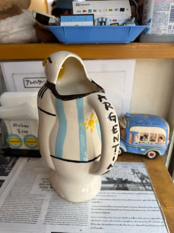 Un cántaro cerámico blanco con franjas celestes, blancas y un sol amarillo, y la palabra "ARGENTINA", junto a una miniatura de un autobús azul