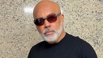 Hombre calvo con barba canosa, gafas de sol y camiseta negra delante de una pared de granito