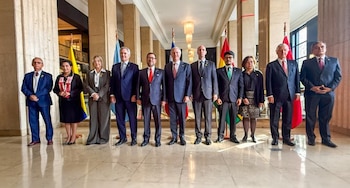 Once funcionarios, hombres y mujeres, posan formalmente de pie en un elegante salón con columnas y suelo de mármol, frente a banderas de países sudamericanos
