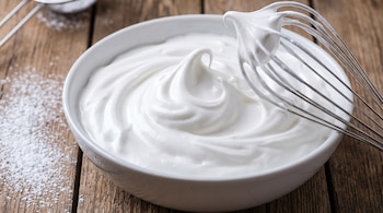 Un bol blanco lleno de merengue suizo, un batidor de metal con merengue, azúcar en polvo esparcida y una mesa de madera.