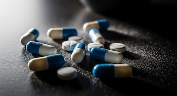 Varias pastillas blancas y cápsulas azules y blancas esparcidas sobre una superficie oscura. Se observa polvo fino alrededor de las pastillas con luz dramática.