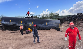Varios vagones cisterna de tren negros descarrilados sobre tierra. Cuatro trabajadores con cascos y ropa de seguridad están en el sitio. Cielo azul con nubes