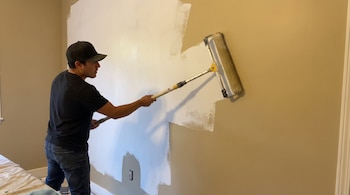 Un hombre de espaldas y perfil usa un rodillo de extensión para pintar una pared beige de blanco. Parte de la pared ya está pintada. Se ve una toma de corriente.
