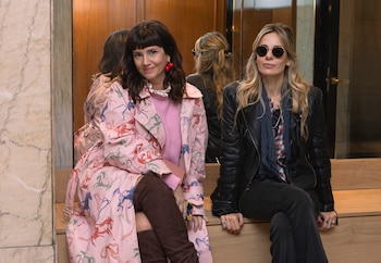 Dos mujeres sentadas en un banco de madera: una con abrigo rosa de caballos, aretes de cereza; la otra con chaqueta de cuero negra y gafas de sol