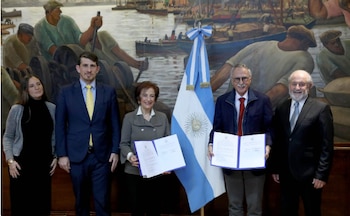 Cinco personas de pie frente a una bandera de Argentina y un mural. Dos de ellas, una mujer y un hombre, sostienen documentos