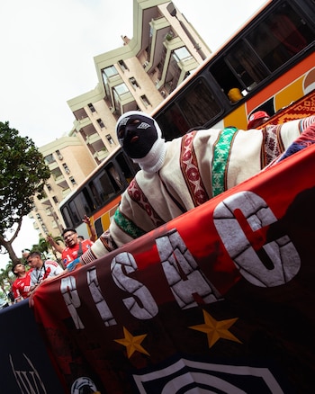 Un aficionado encapuchado con poncho andino sostiene una banderola roja junto a un autobús y otros hinchas vestidos con camisetas rojas