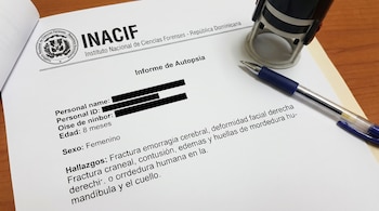 Un documento oficial del INACIF titulado 'Informe de Autopsia' sobre una bebé de 8 meses, con hallazgos médicos detallados, yace sobre una mesa de madera junto a un bolígrafo azul y un sello.