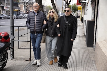 Tres personas caminan en una acera urbana. Una mujer de cabello rubio con gafas de sol va entre dos hombres. Hay autos estacionados y un poste de luz