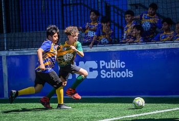 Dos jóvenes en uniformes de fútbol corren en un campo de césped artificial con un balón. Niños observan detrás de una red y muro azul