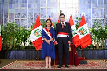 Mujer con vestido azul y banda presidencial, hombre con traje oscuro y banda presidencial, varias banderas de Perú, alfombra roja, plantas, vitrales
