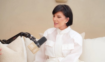 Retrato de Kris Jenner, una mujer de cabello oscuro y corto, vestida con una camisa blanca, sentada junto a un micrófono en un sofá claro