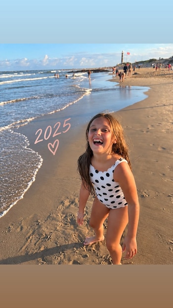 Una niña con traje de baño blanco con lunares negros sonríe en la arena junto al agua en una playa