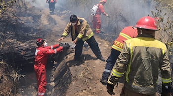 Bomberos con uniformes de protección, cascos y mascarillas pasan un bidón en un terreno de tierra con humo y vegetación quemada.
