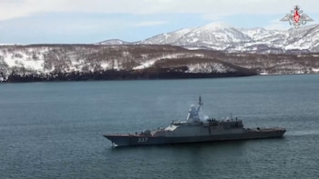 Vista aérea de un buque de guerra gris con el número 337 en el casco, navegando en aguas azules. El fondo muestra montañas cubiertas de nieve y árboles