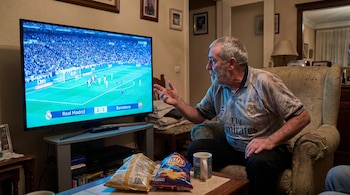 Un hombre de edad avanzada con barba, vistiendo una camiseta del Real Madrid, mira un partido de fútbol en un televisor grande en su sala, gesticulando con una mano.