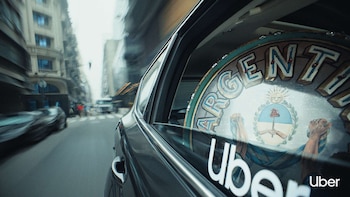 Exterior de coche oscuro con fondo de calle urbana borrosa. Ventana trasera muestra un cartel redondo con parte de "ARGENTINA", escudo nacional y logo Uber