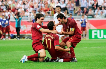 Tres futbolistas de Portugal con camisetas rojas celebran un gol en el campo. Cristiano Ronaldo (17) de rodillas, abrazado por Luis Figo y otro jugador, con público al fondo